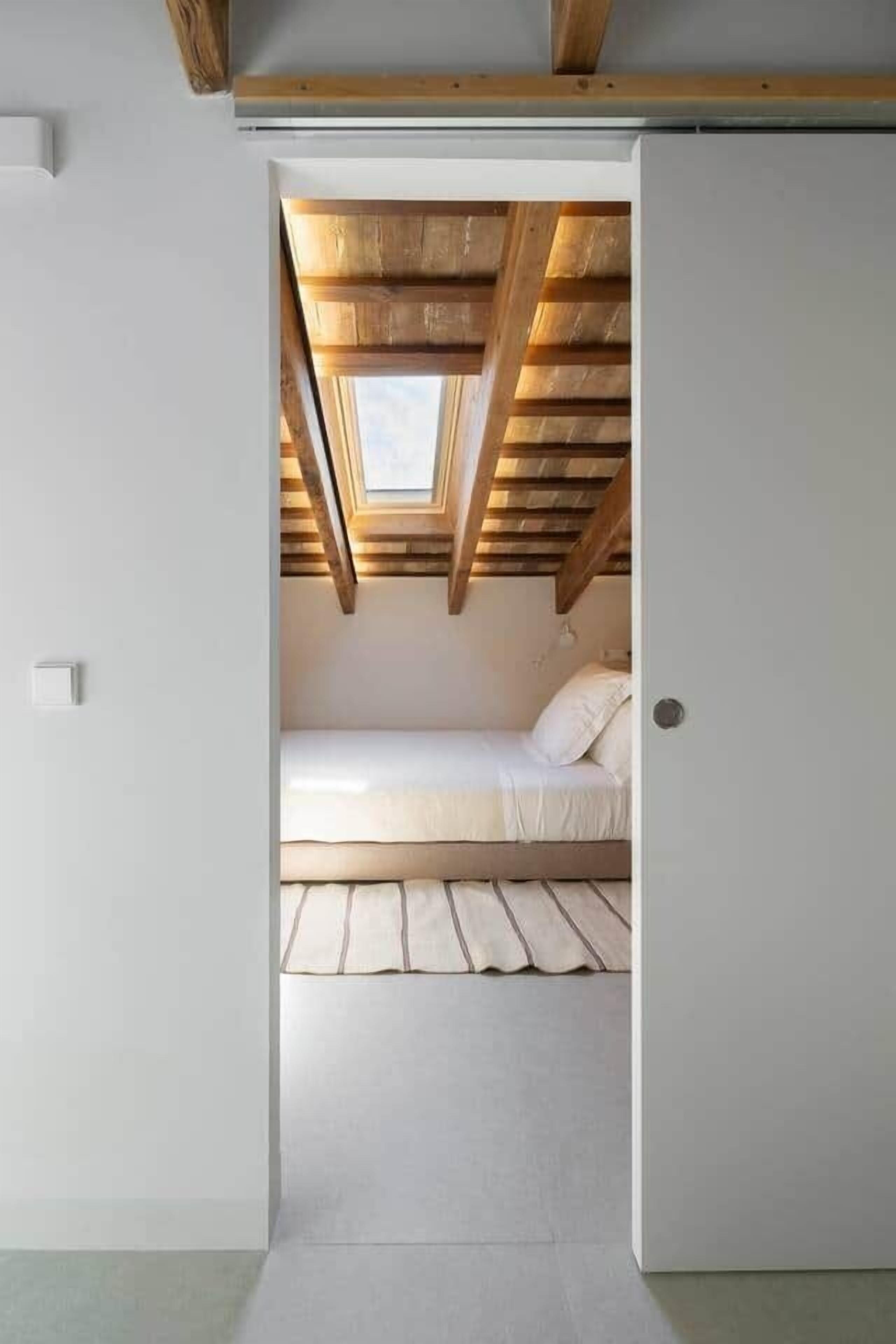 A narrow doorway leading to a loft bedroom with a skylight, a bed, and wooden beams.