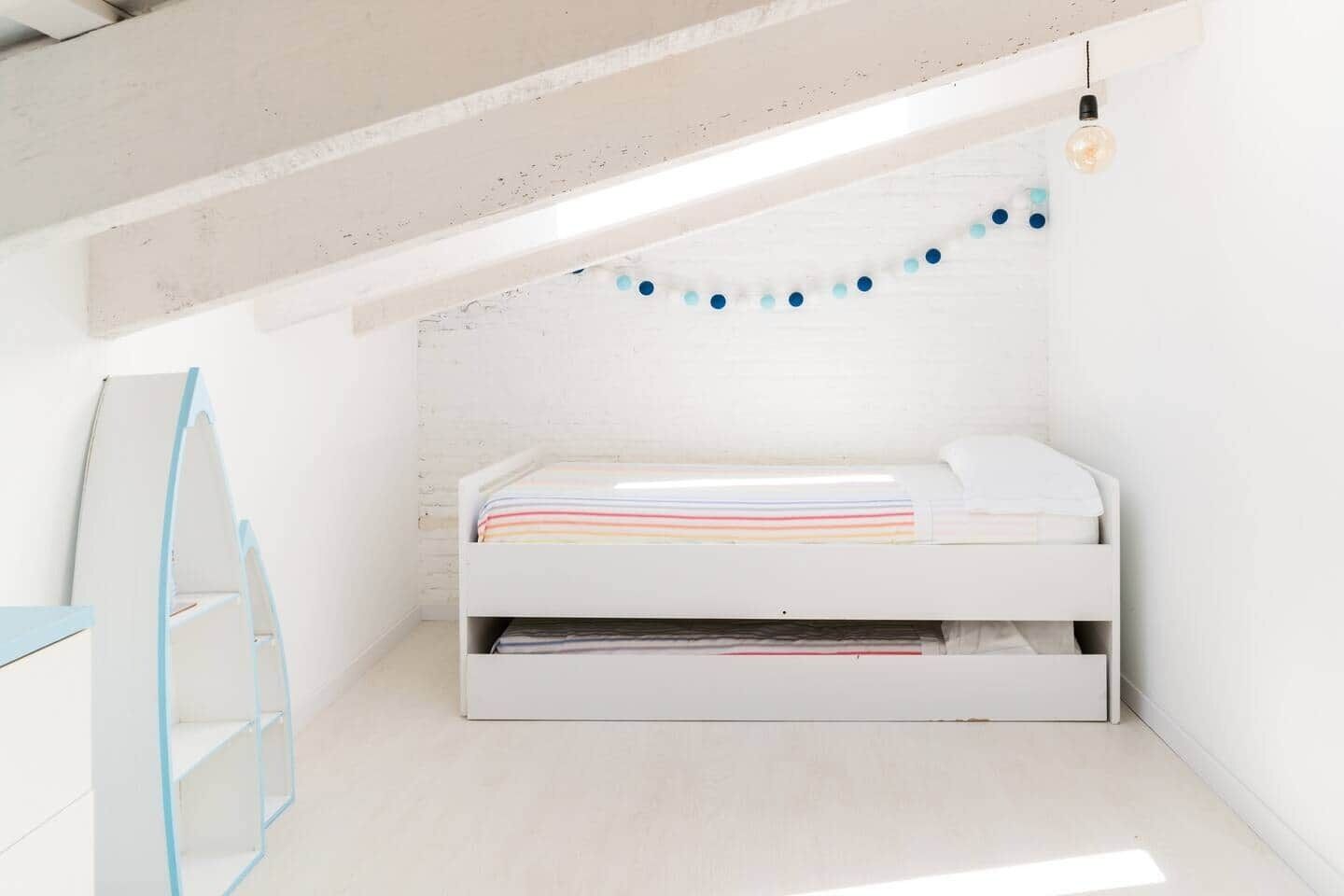Un dormitorio estilo loft blanco con una cama nido, una escalera y una guirnalda de luces azules.