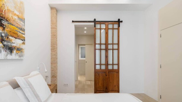 Un dormitorio con cama, puerta corredera de madera, pared blanca y un cuadro.