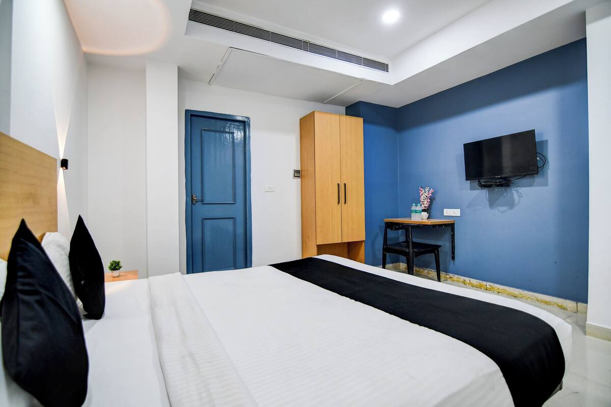 A bedroom with a bed, a blue door, a wooden wardrobe, a small table with a TV, and a wall-mounted TV.