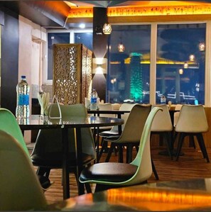 Un restaurante con vista a un paisaje urbano nocturno, muebles modernos y una pared decorativa.
