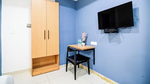 A room with a wooden wardrobe, a small table with a chair, a flat-screen TV mounted on the wall, and a power outlet on the wall.