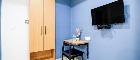 A room with a wooden wardrobe, a small table with a chair, a flat-screen TV mounted on the wall, and a power outlet on the wall.