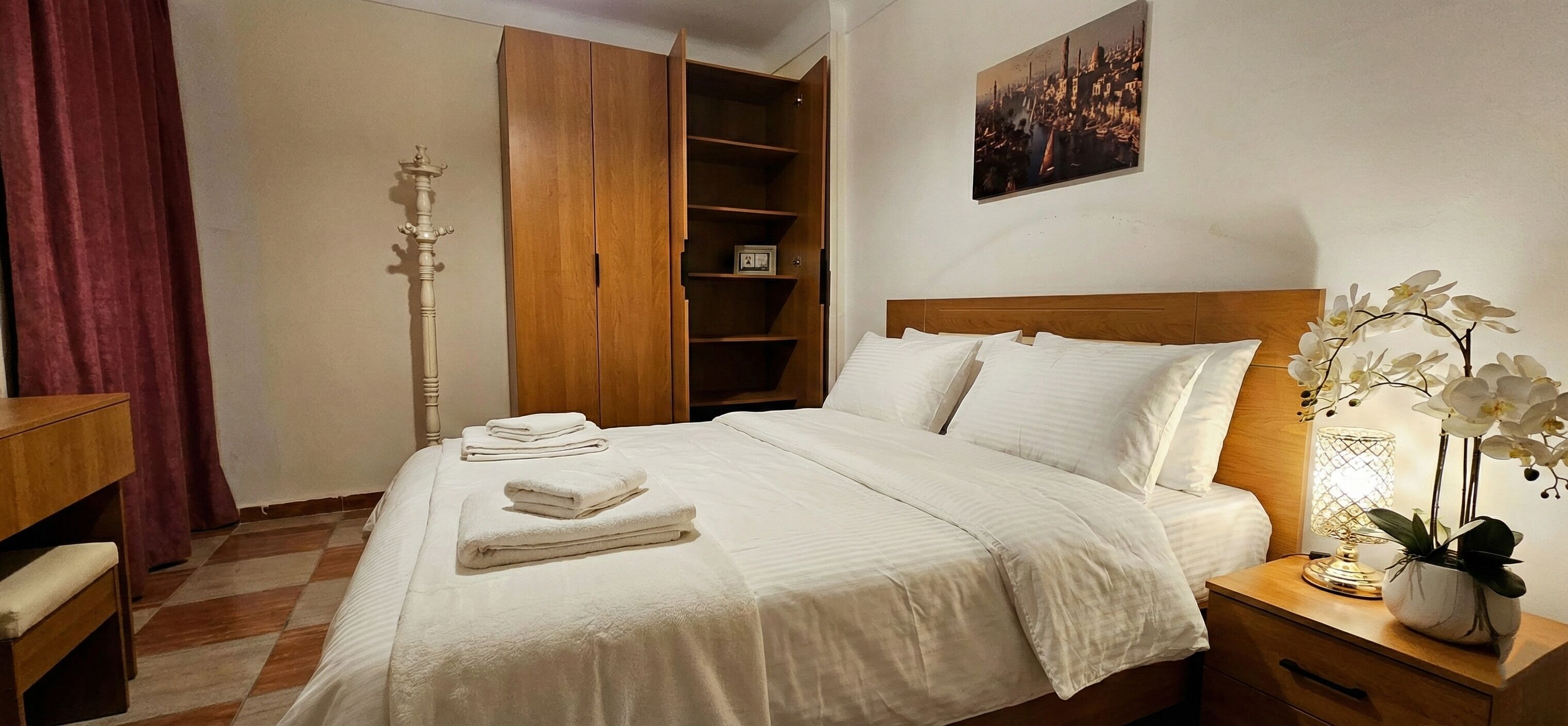 A neatly made bed with white linens and towels, a wooden headboard, a bedside lamp, and a nightstand with a potted orchid.