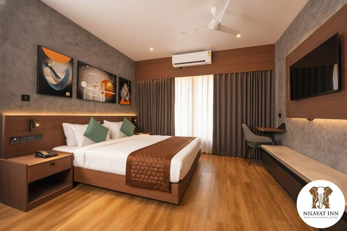 A modern hotel room with a large bed, a desk, and a TV. There is an air conditioning unit on the wall and a ceiling fan.