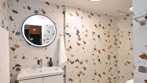 Un baño con lavamanos blanco, espejo redondo y zona de ducha con suelo de terrazo.