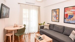 Una sala moderna con un sofá gris, una mesita redonda y una mesa de centro. Hay un televisor montado en la pared y una ventana con cortinas.
