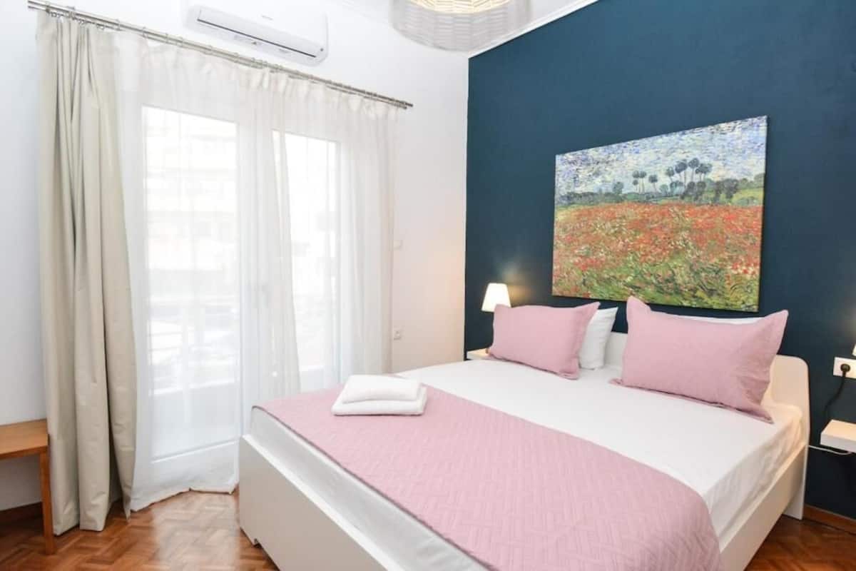 A bedroom with a bed, white curtains, a painting, and a bedside lamp.