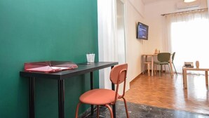 Una habitación con pared verde, una mesa negra con una carpeta roja, una taza blanca y una silla. Hay un televisor en la pared y una ventana con cortinas.