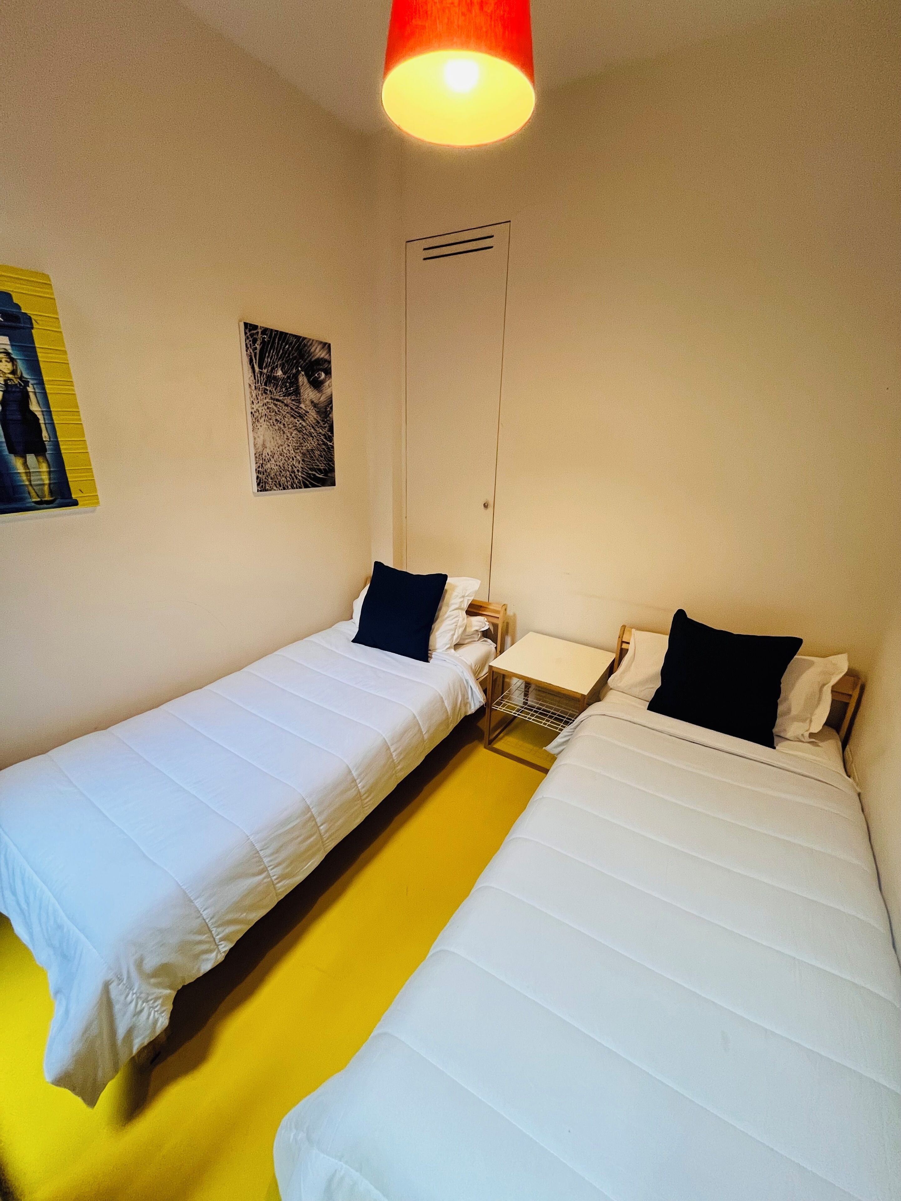 A room with two single beds, a small table, a wall-mounted shelf, and a ceiling light.