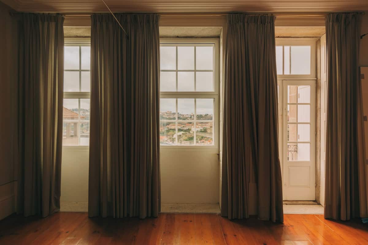 A room with wooden flooring, a large window with curtains, and a door leading to another room.