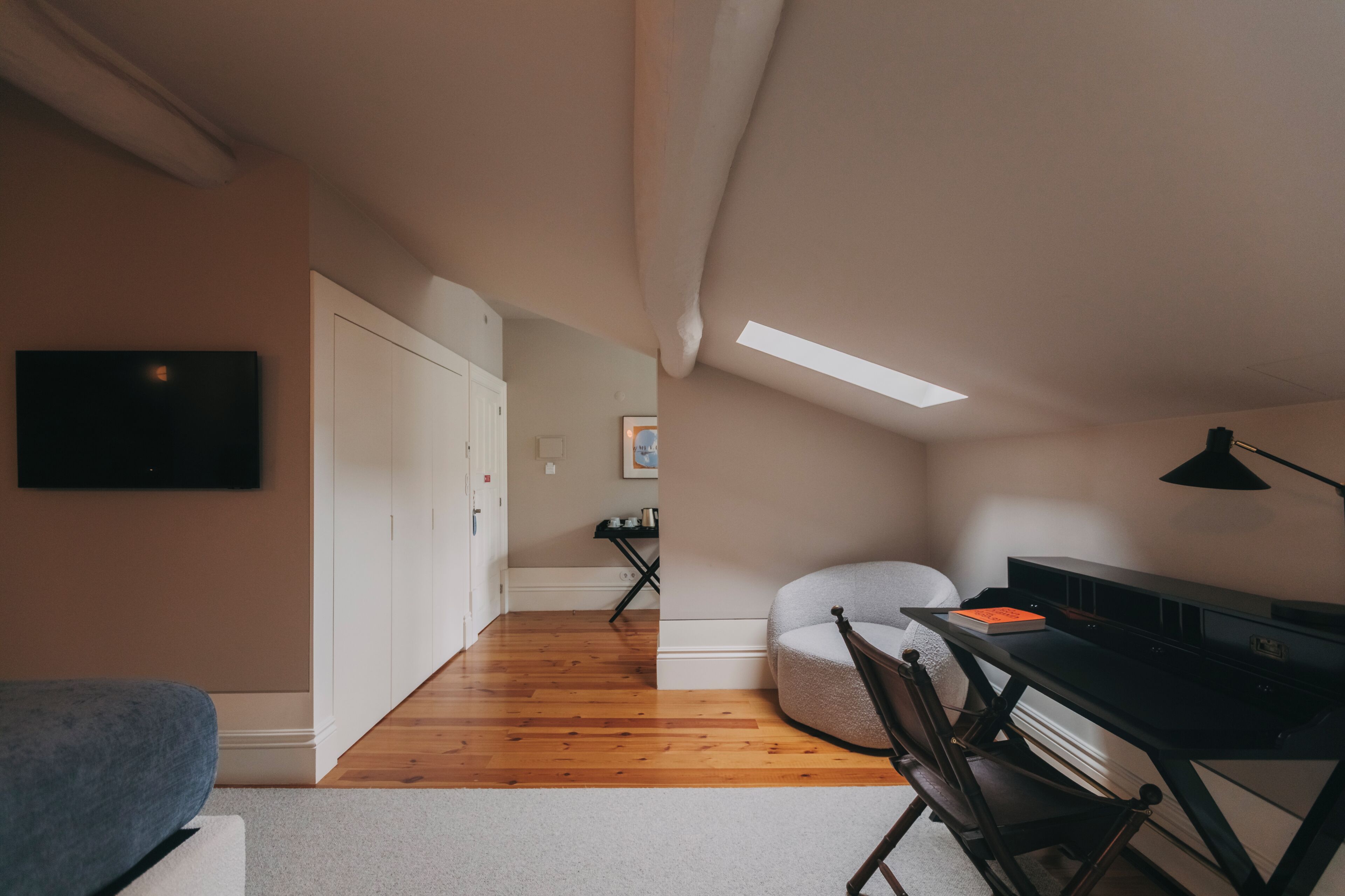 A modern, compact living space with a wooden floor, a sofa, a desk with a chair, and a TV mounted on the wall.