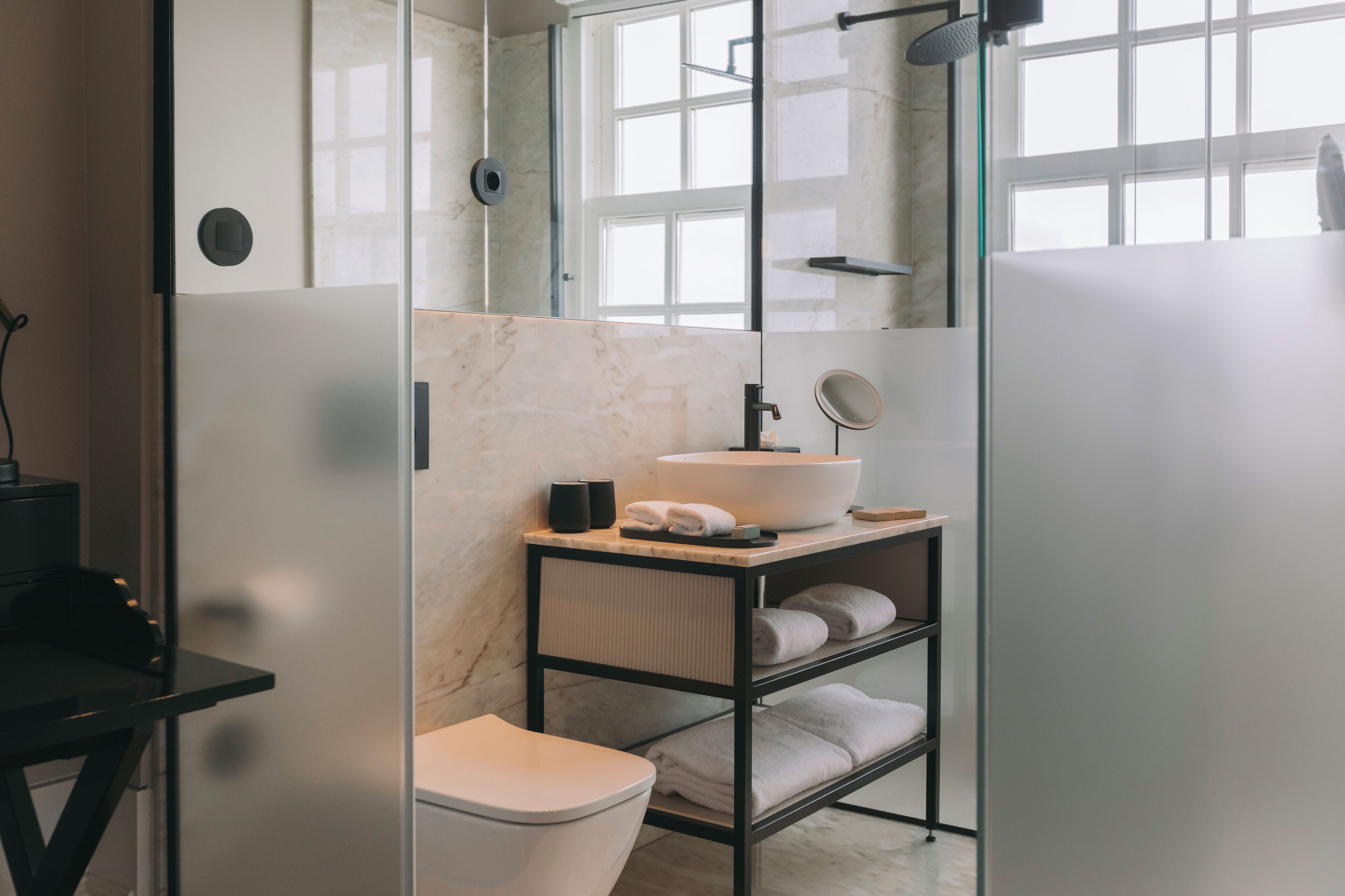 A modern bathroom with a glass shower enclosure, a white sink, and a black shelf with towels.