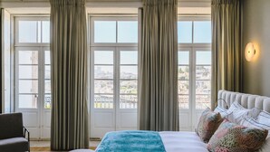 A hotel room with a large bed, two pillows, a chair, and a view of a cityscape through large windows with curtains. - Torel Private Collection (Porto)
