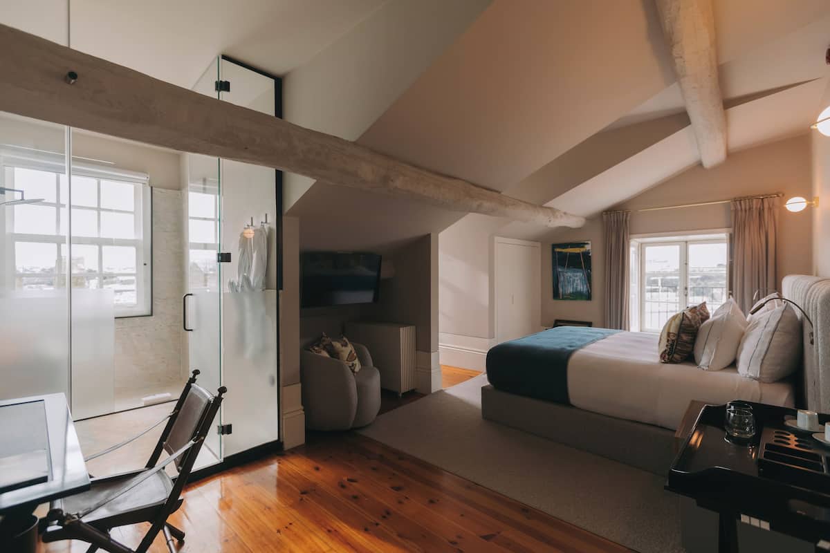 A modern bedroom with a large bed, a desk, and a chair. There is a window with curtains, a radiator, and a TV mounted on the wall.