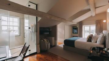 Ein modernes Schlafzimmer mit einem großen Bett, einem Schreibtisch und einem Sessel. Ein Fenster mit Vorhängen, eine Heizung und ein an der Wand befestigtes Fernsehgerät sind ebenfalls vorhanden.