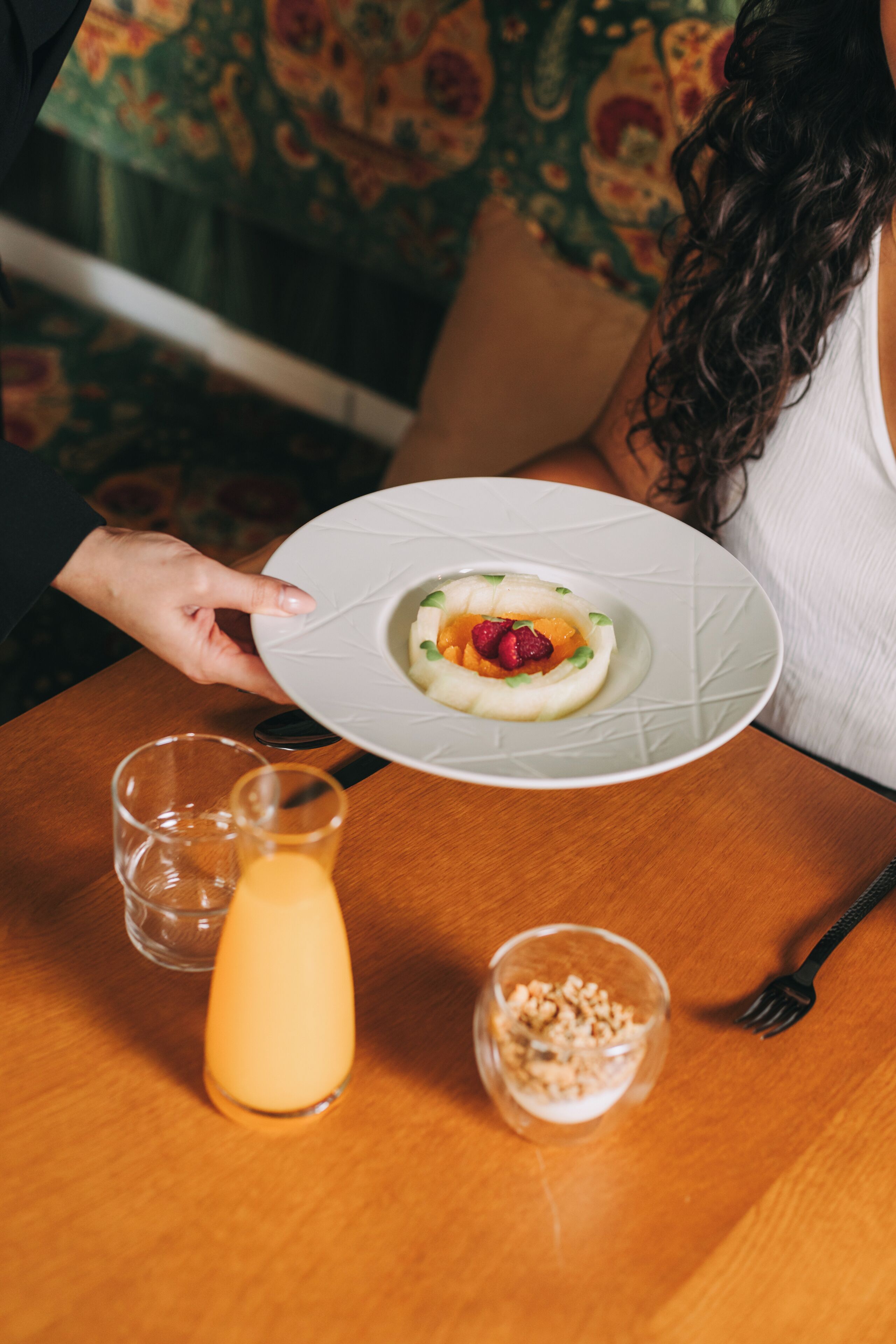A person holding a plate with a dessert, a glass of orange juice, and a small bowl of granola on a wooden table.