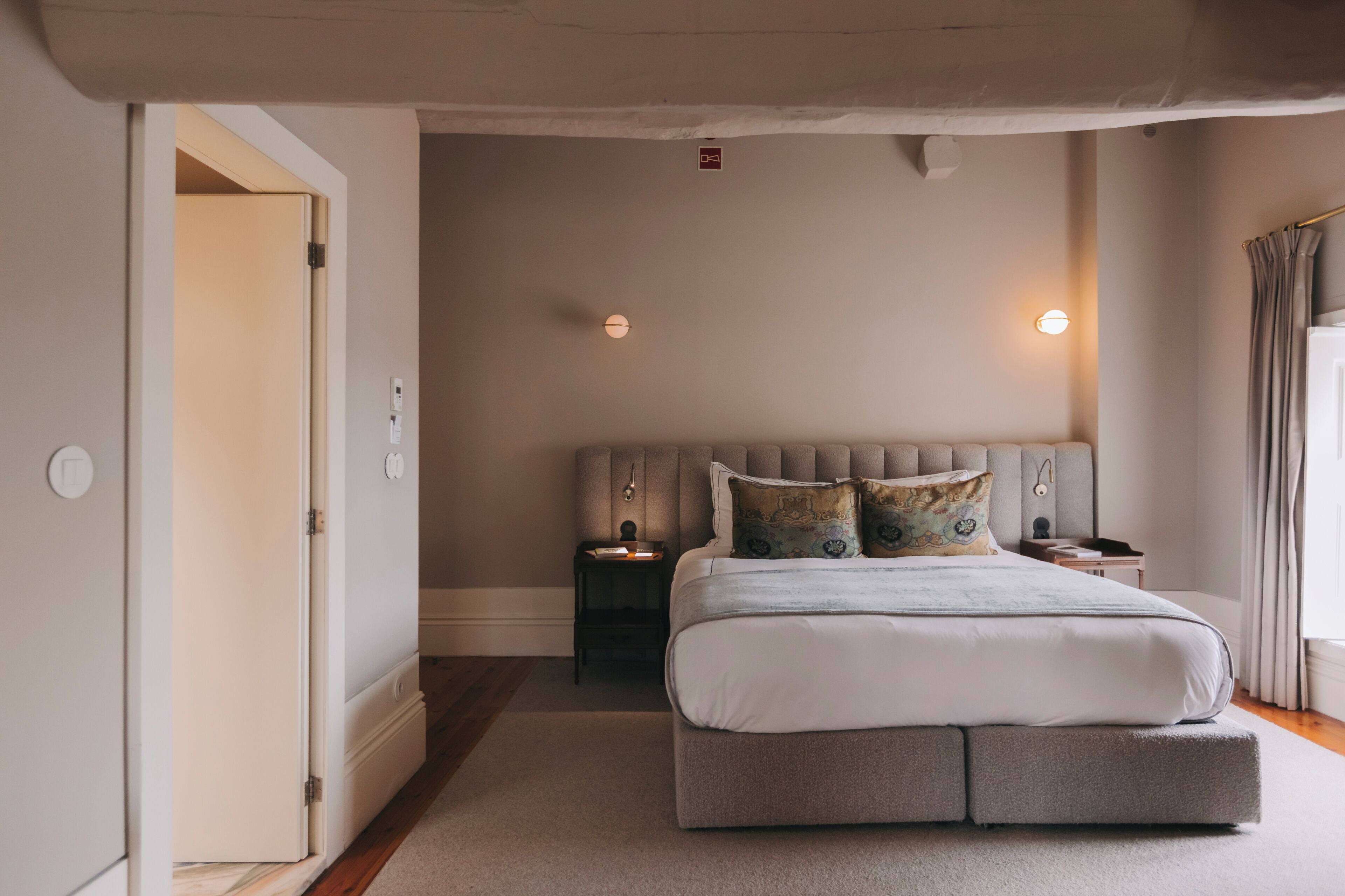 Un dormitorio con una cama, mesitas de noche, una lámpara de pared y una puerta.