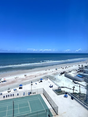 Blick auf einen Strand mit Tennisplatz, Sonnenschirmen und Liegestühlen.