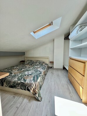 Ein Schlafzimmer mit schrägen Wänden, einem Bett mit blattgemustertem Überwurf, einer Holzkommode und einem Oberlicht.
