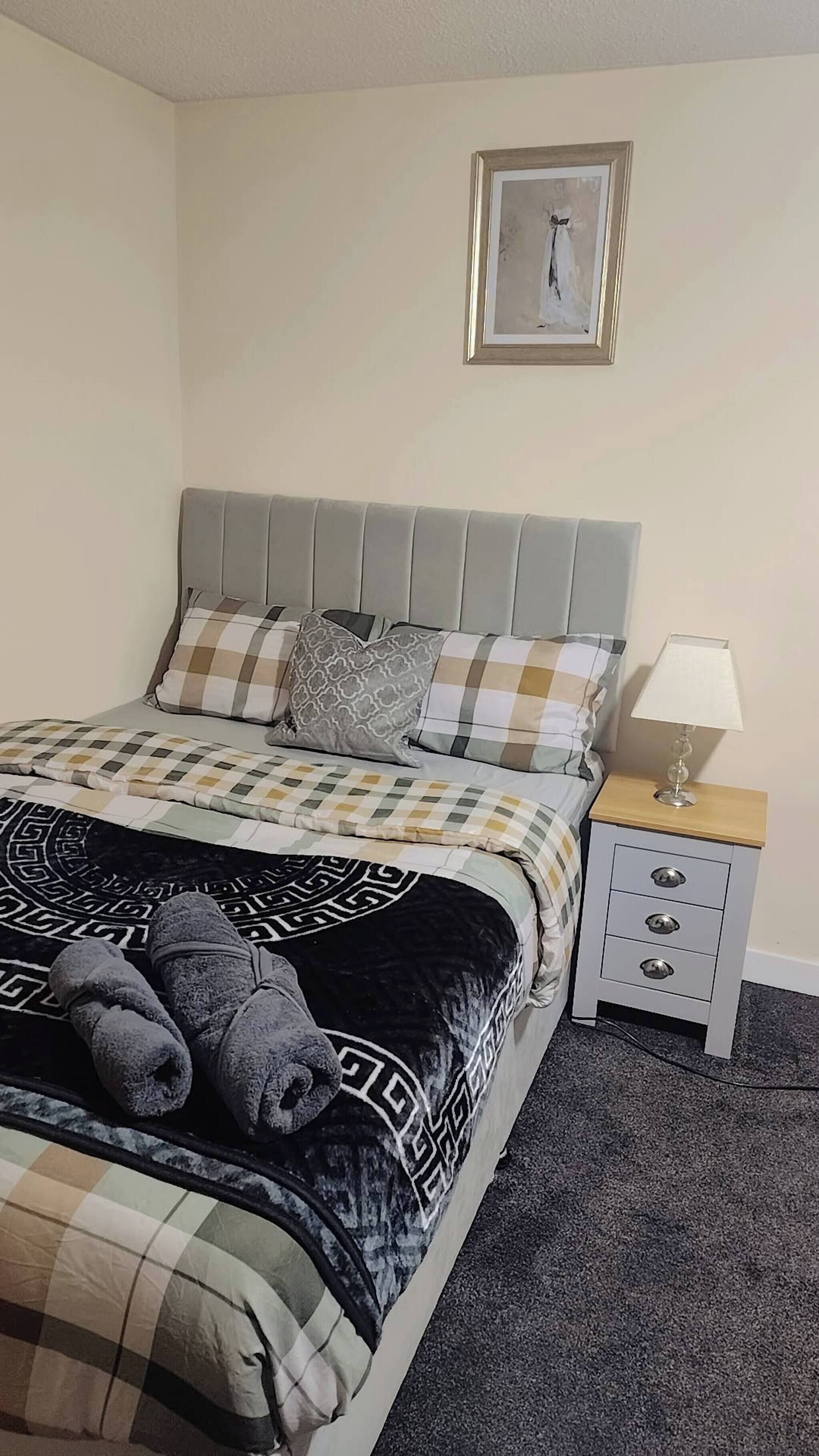A neatly made bed with a checkered duvet, pillows, and a blanket with a geometric pattern. There is a bedside table with a lamp and a small drawer unit. A framed picture hangs on the wall above the bed.
