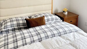 A bed with a headboard, a nightstand with a vase of flowers, and a plain wall.
