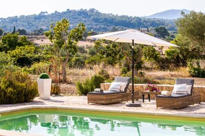 Un espace aménagé au bord de la piscine, avec des fauteuils de relaxation en osier, un grand parasol et une vue sur un paysage montagneux.