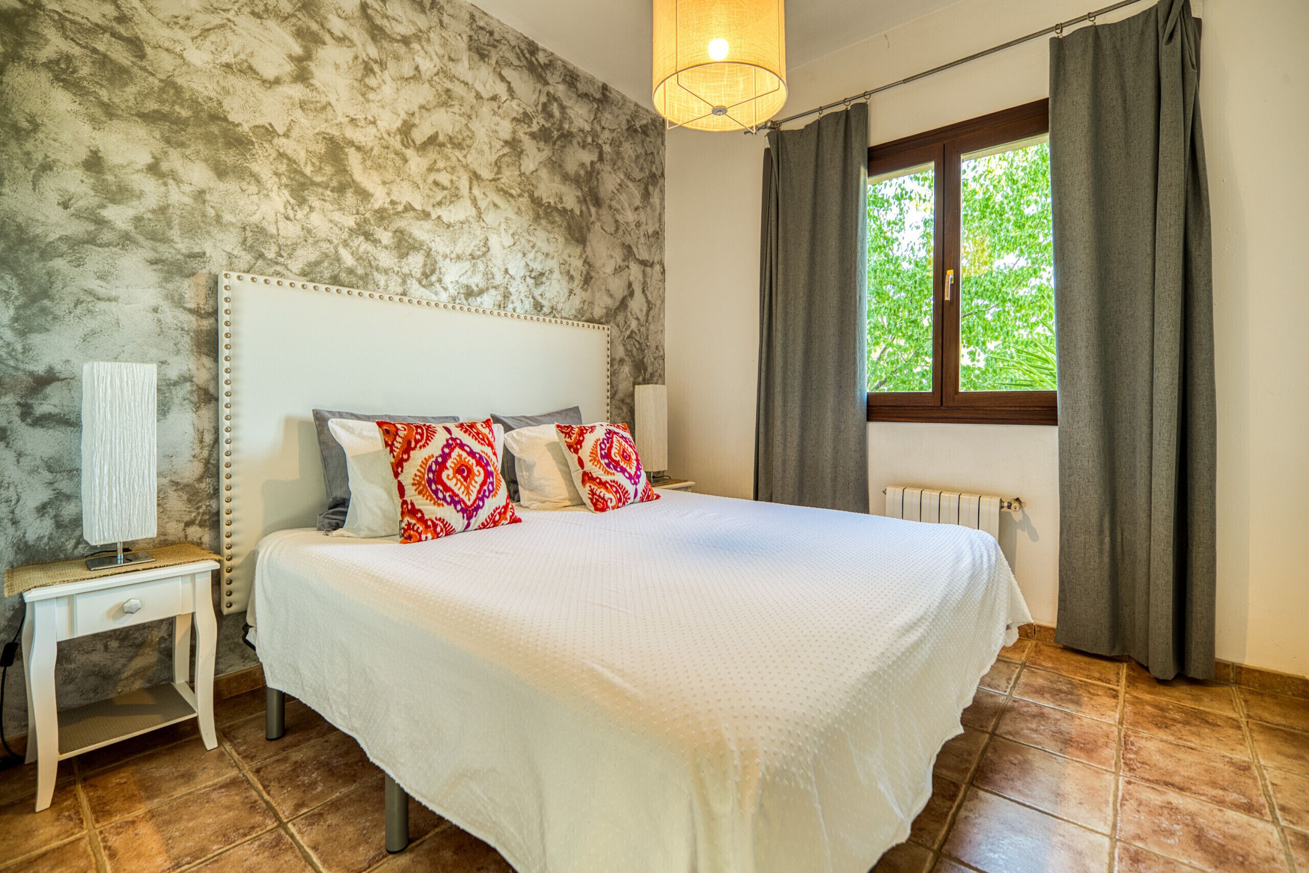 A bedroom with a large bed, a nightstand, a lamp, a window with curtains, and a radiator.