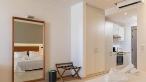 A hotel room with a bed, a mirror, a small wooden bench, a trash bin, and a kitchenette. - Mini Villa with Pool View (Malia)