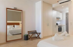 Une chambre d’hôtel comprenant un lit, un miroir, un étendoir en bois, une poubelle et une kitchenette.