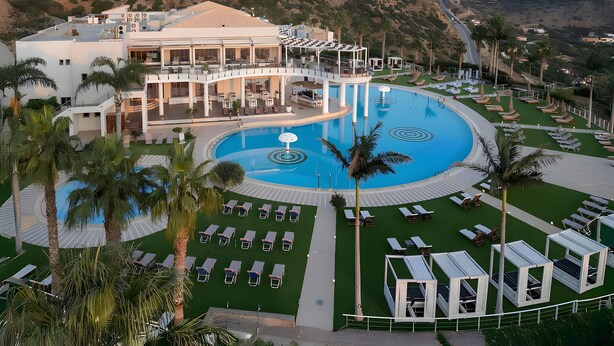 Ett semesterhotell med en stor pool i mitten, flera avslappningsområden och en byggnad i flera våningar med sittplatser utomhus.