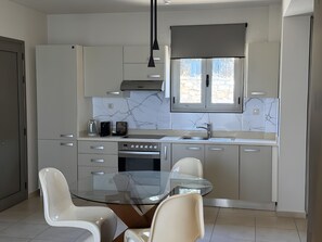 A modern kitchen with a glass dining table, white cabinets, and a built-in oven. - Villa 'One Bedroom Villa with Pool View' with Wi-Fi and Air Conditioning (Malia)