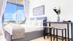 A modern hotel room with a bed, a desk with two chairs, a large window with curtains, and a framed picture on the wall. - Bloomfields Stu Riviera Skies (Dubai)