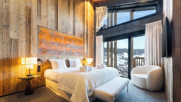 Une chambre d’hôtel avec un grand lit, un coin salon avec un fauteuil, une vue sur un paysage enneigé par de grandes fenêtres et un mur en bois orné d’un tableau.