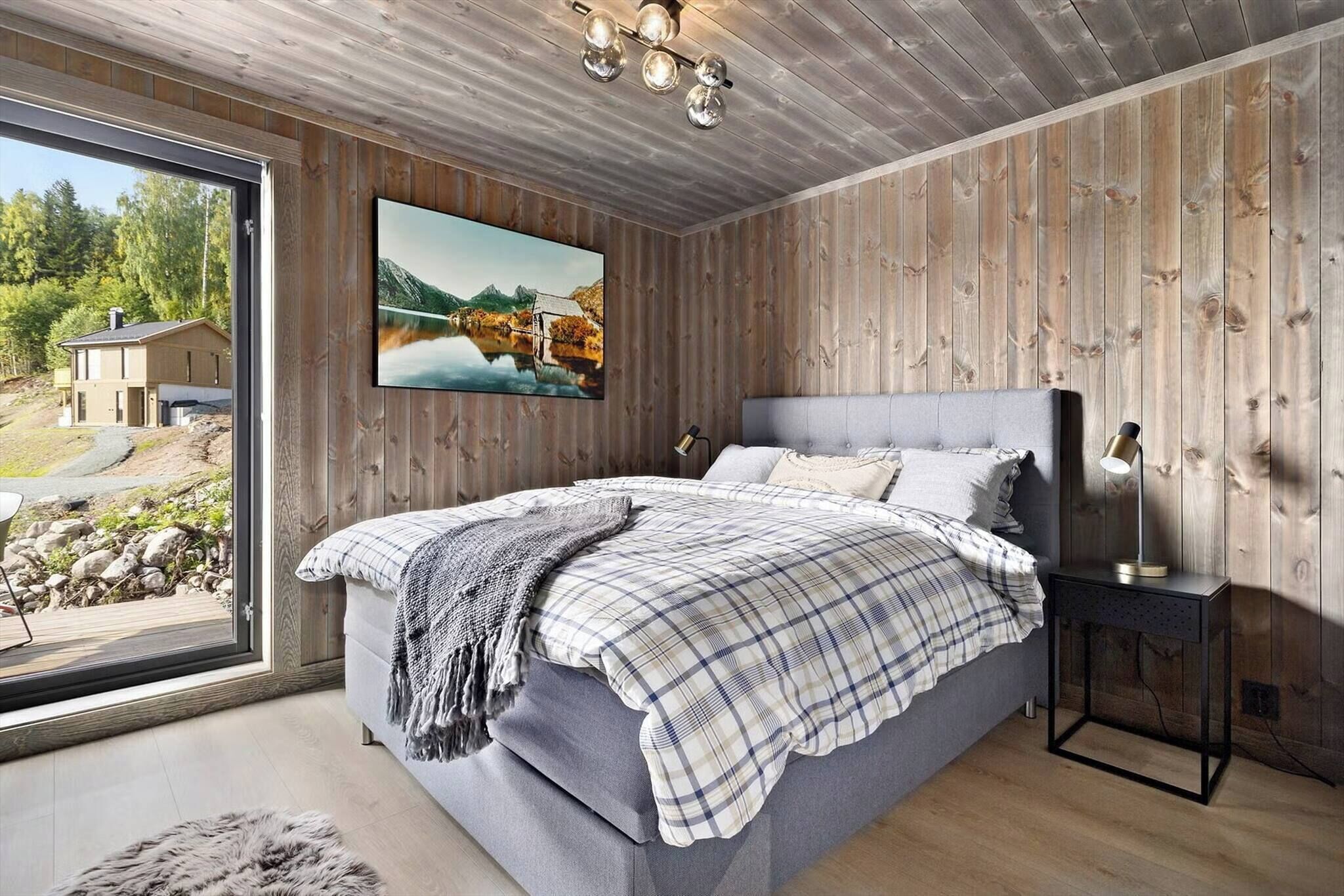 A bedroom with a large bed, a wooden wall, a painting, and a view of a house and landscape through a window.