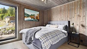 Een slaapkamer met een groot bed, een houten wand, een schilderij en uitzicht op een huis en landschap door een raam.