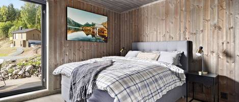 A bedroom with a large bed, a wooden wall, a painting, and a view of a house and landscape through a window.