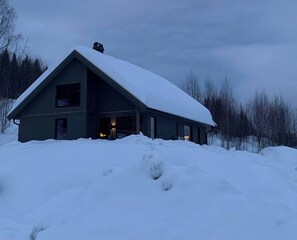 A two-story cabin with a steep roof, surrounded by snow-covered ground and trees.