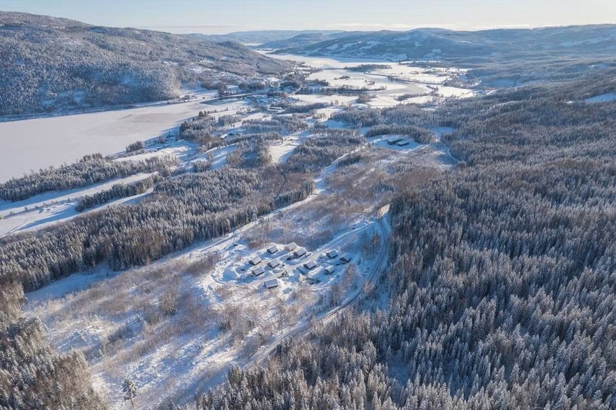 An aerial view of a snow-covered landscape with a cluster of buildings, likely a resort or lodge, surrounded by dense forest and a frozen lake.