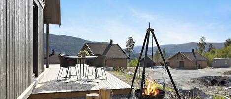 Una terraza de madera con mobiliario exterior, una fogata y vistas de casas y colinas circundantes.