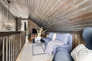 A cozy loft-style living area with a wooden ceiling, a blue sofa, a coffee table, and a TV in the background. - New and Great Cabin at the Foot of Norefjell (Krodsherad)