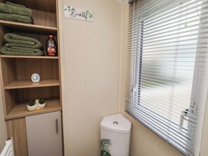 A compact bathroom with a toilet, a shelf with folded towels, a small decorative item, and a window with blinds. - Homeslea 33 Millennium Gardens (Morpeth)