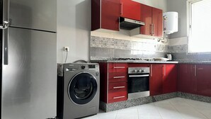 Une cuisine compacte équipée d’un réfrigérateur en acier inoxydable, d’un lave-linge, de meubles de rangement rouges, d’un four encastré et d’un chauffe-eau.