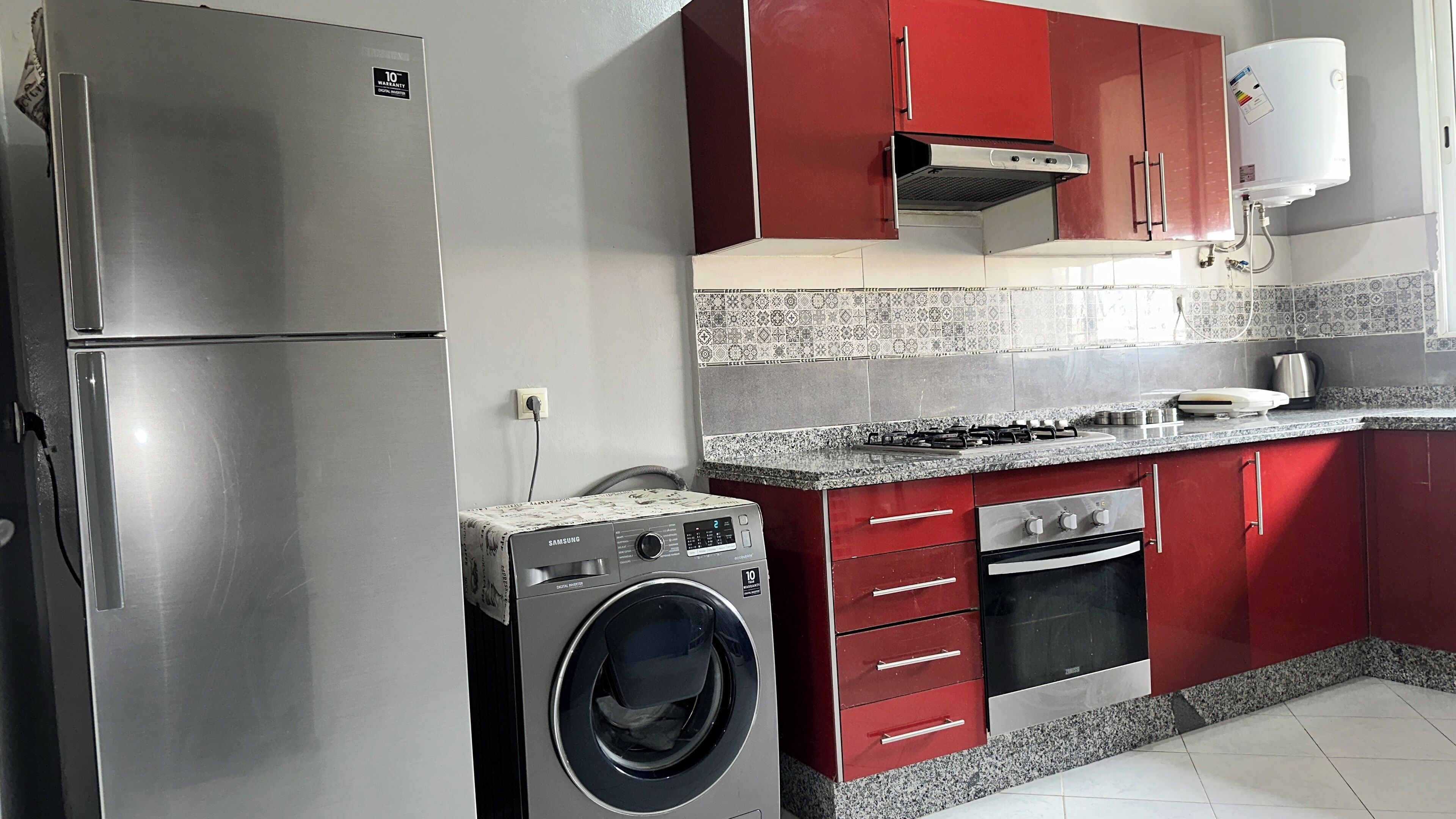Een compacte keuken met een roestvrijstalen koelkast, een wasmachine, een gasfornuis en rode keukenkastjes.