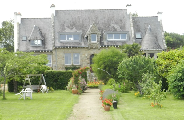 A large stone building with a symmetrical facade, a central archway, and a well-maintained garden with a swing set and potted plants. - An exceptional spot! Apartment in a charming house, swimming at the bottom of the garden (Locmariaquer)