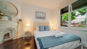 A bedroom with a bed, two bedside tables, a mirror, a fireplace, and a window with a view of greenery. - The Hidden Courtyard | Erskineville (Erskineville)