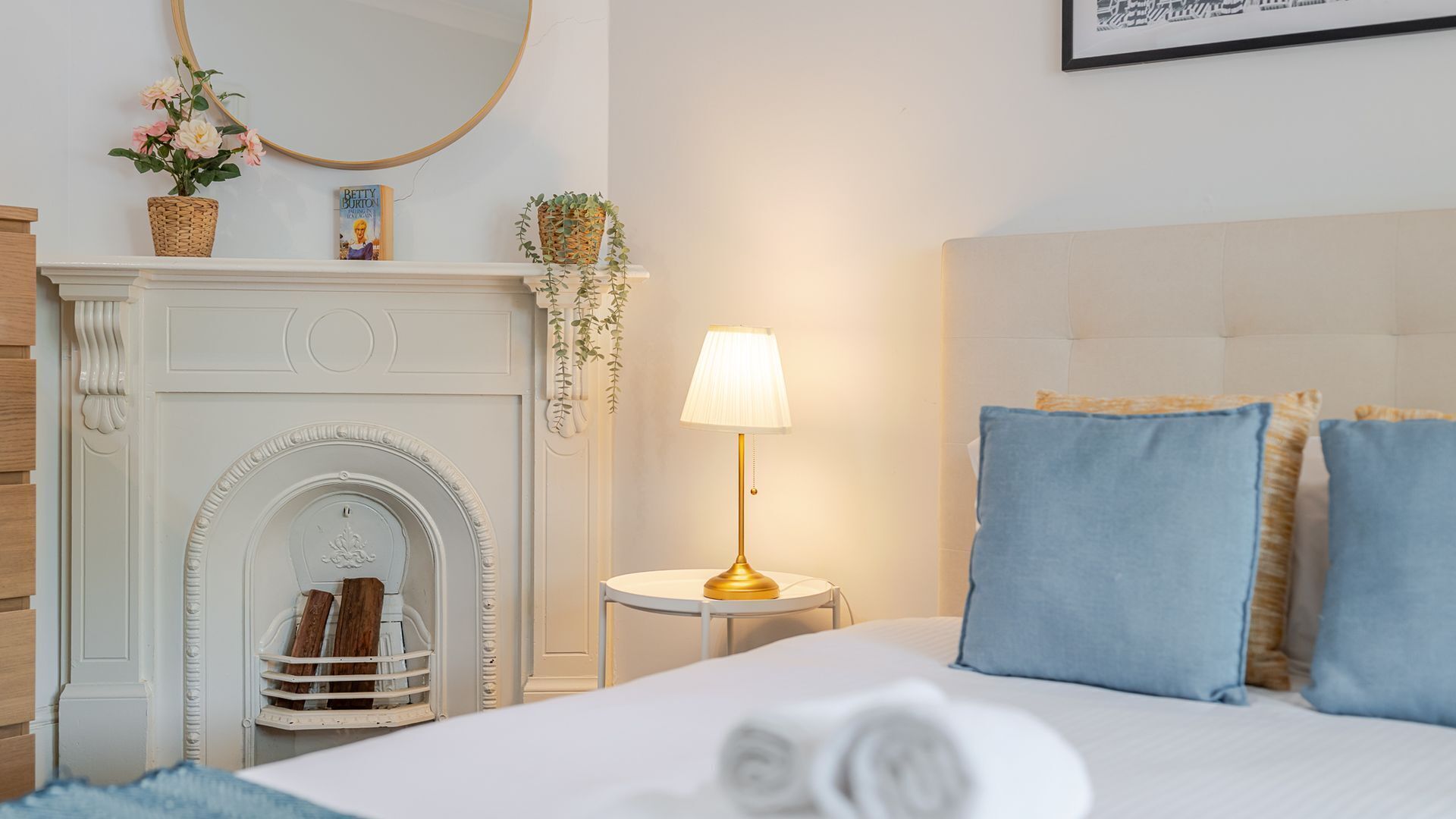 Habitación de hotel con chimenea blanca, espejo redondo, mesita de noche con lámpara y cama con almohadas azules.