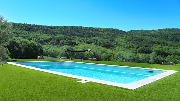 A rectangular swimming pool with a white edge, surrounded by well-manicured green grass, with a hammock and rocks in the background. - Provencal house with heated pool, jacuzzi and view 5 minutes from the Colorado (Viens)