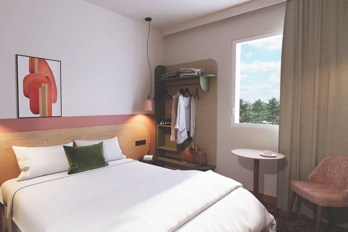 A hotel room with a bed, bedside table, a small round table, a chair, a coat rack, and a window with a view.