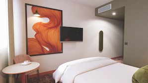 A hotel room with a bed, a television mounted on the wall, a small table with a cup, and a painting.